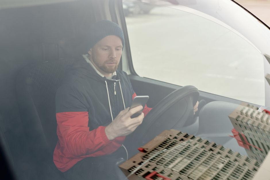 A person wearing a white short-sleeved shirt is inside a vehicle, holding a pen and writing on a clipboard that is resting on a cardboard box. Their left hand is gripping the steering wheel, indicatin