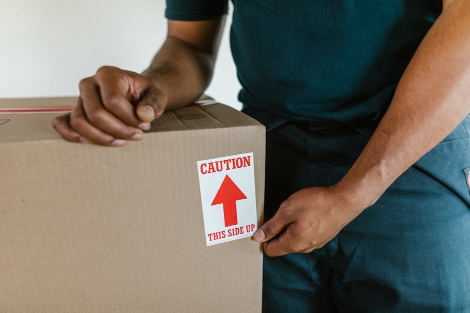 A young man with long hair, wearing a black bandana and a dark blue shirt, is sitting in the driver's seat of a white moving van with the words 'MOVING COMPANY LOCAL & LONG DISTANCE' printed on the si