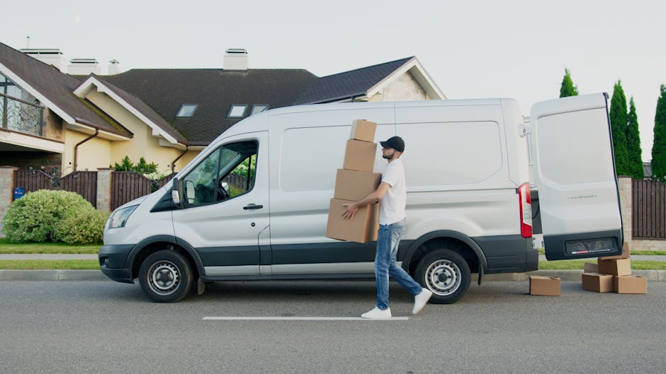 A man with dark hair and a beard, dressed in a white t-shirt, blue jeans, and white sneakers, is carrying three stacked cardboard boxes outside a white commercial van parked on a suburban street. He is walking toward the van's open rear cargo area, which contains additional cardboard boxes and packing materials. The van is positioned on the road in front of a residential property with a modern house featuring a dark roof, light-colored walls, a balcony with a glass railing, and well-maintained greenery including bushes and tall trees. Several more cardboard boxes are on the pavement nearby, some placed on the ground, indicating the process of home relocation, furniture transport, or packing and moving. The scene captures the loading process typical of house removals services such as those offered by Man with Van Sutton, with the van aligned for efficient access in a narrow access area, highlighting the logistical considerations involved in house removals with limited space.