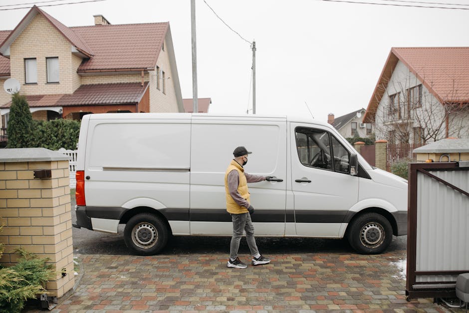 A man wearing a face mask, glasses, a beige and brown jacket, dark trousers, and sneakers is standing on a brick driveway in front of a residential property, holding the door handle of a white panel van. The van is parked parallel to the curb, partially blocking the view of the house behind it. The house features a beige brick exterior, a pitched roof with red tiles, and multiple windows. The driveway is paved with multicoloured bricks, and there are brick and metal boundary walls on either side of the driveway. In the background, neighboring houses with similar pitched roofs and red tiles, utility poles, and power lines are visible. The scene appears to be part of home relocation or furniture transport activities, with the individual possibly preparing to load or unload items as part of a house removal service, typical of a professional moving process conducted by Man with Van Sutton.
