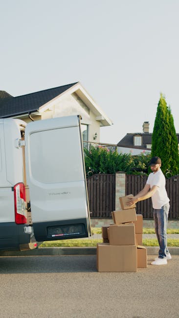 A man with dark hair and a beard, dressed in a white t-shirt, blue jeans, and white sneakers, is carrying three stacked cardboard boxes outside a white commercial van parked on a suburban street. He is walking toward the van's open rear cargo area, which contains additional cardboard boxes and packing materials. The van is positioned on the road in front of a residential property with a modern house featuring a dark roof, light-colored walls, a balcony with a glass railing, and well-maintained greenery including bushes and tall trees. Several more cardboard boxes are on the pavement nearby, some placed on the ground, indicating the process of home relocation, furniture transport, or packing and moving. The scene captures the loading process typical of house removals services such as those offered by Man with Van Sutton, with the van aligned for efficient access in a narrow access area, highlighting the logistical considerations involved in house removals with limited space.