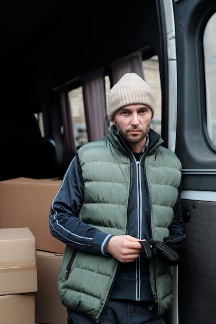 A man wearing a dark jacket and cap is unloading cardboard boxes from the rear of a large white commercial van parked outside a modern building with glass windows and metal walls. The van's rear doors